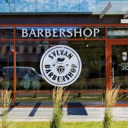 Sylvan Barbershop - Fasada salonu barberskiego z dużym oknem i logo Sylvan Barbershop. Widoczne godziny otwarcia i flaga barberska. W tle odbicie ulicy i nieba.