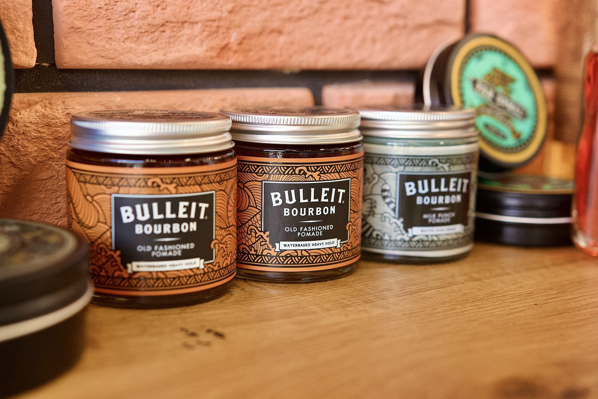 Trzy słoiczki pomady do włosów Bulleit Bourbon na drewnianym blacie, z ceglaną ścianą w tle. Różne warianty produktu, widoczne detale etykiet.