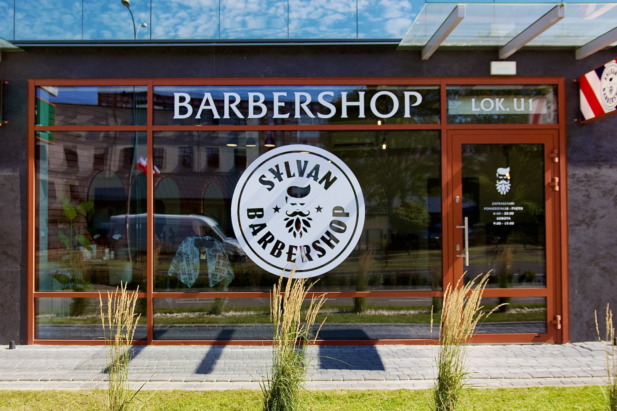Fasada salonu barberskiego z dużym oknem i logo Sylvan Barbershop. Widoczne godziny otwarcia i flaga barberska. W tle odbicie ulicy i nieba.