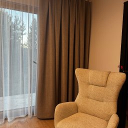Małgorzata Guzik Architektura - Beżowe zasłony zaciemniające i firanka w oknie, obok fotel w Rupniowie. Widok na drzewa za oknem. Przytulne wnętrze.