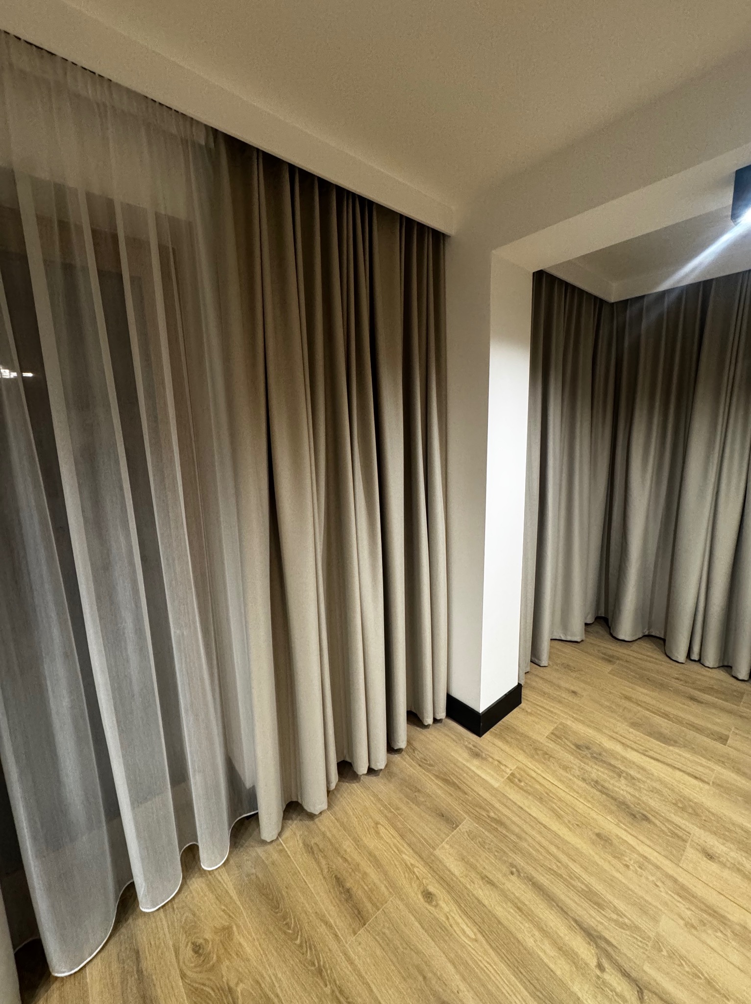 Beżowe, długie zasłony z woalu w jasnym wnętrzu z drewnianą podłogą. Dwie warstwy materiału tworzą efektowne, zaciemniające okrycie okienne.