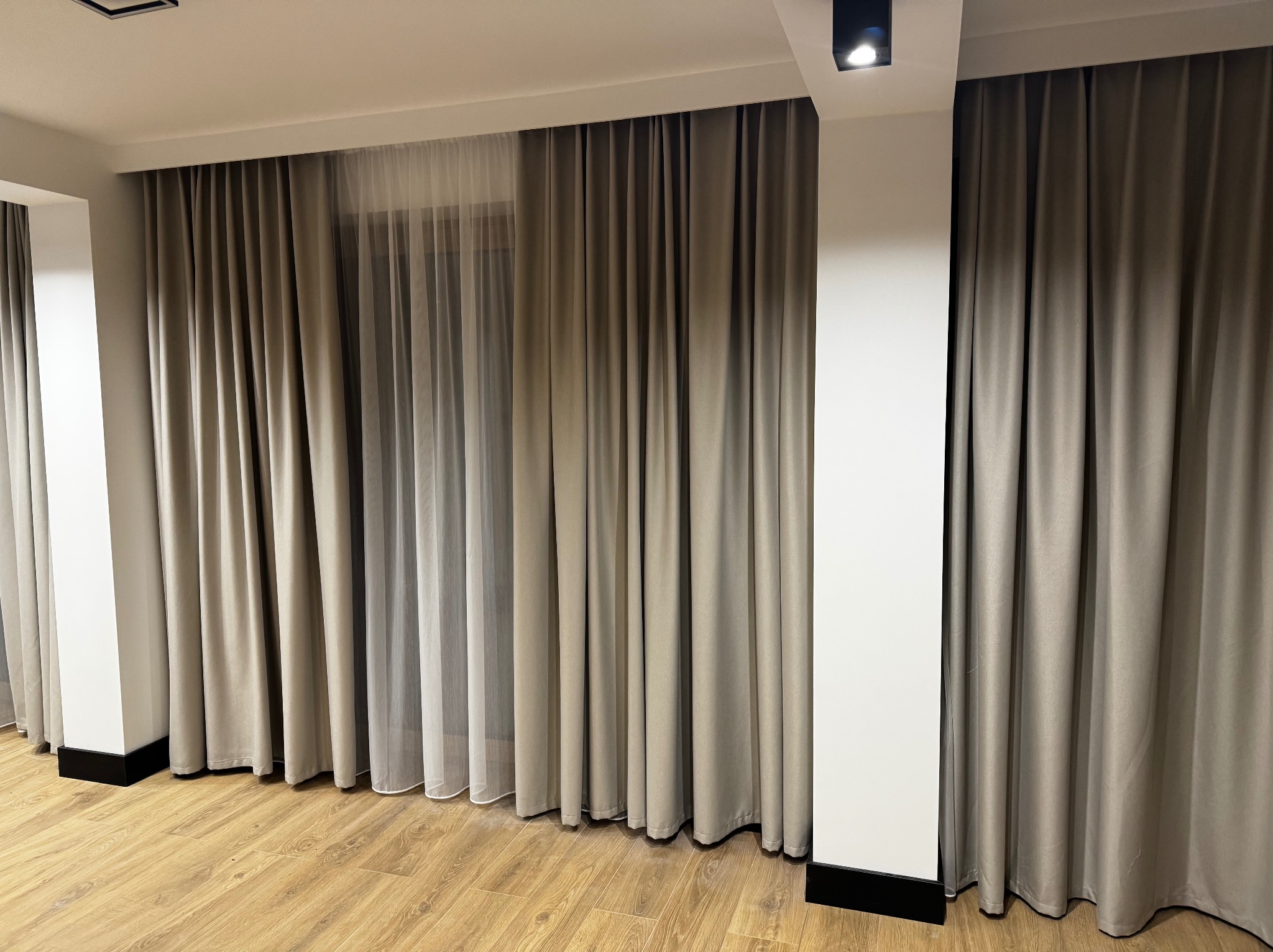 Beżowe, grube zasłony z delikatną, prześwitującą firanką w tle, zawieszone w nowoczesnym wnętrzu z drewnianą podłogą i białymi kolumnami.
