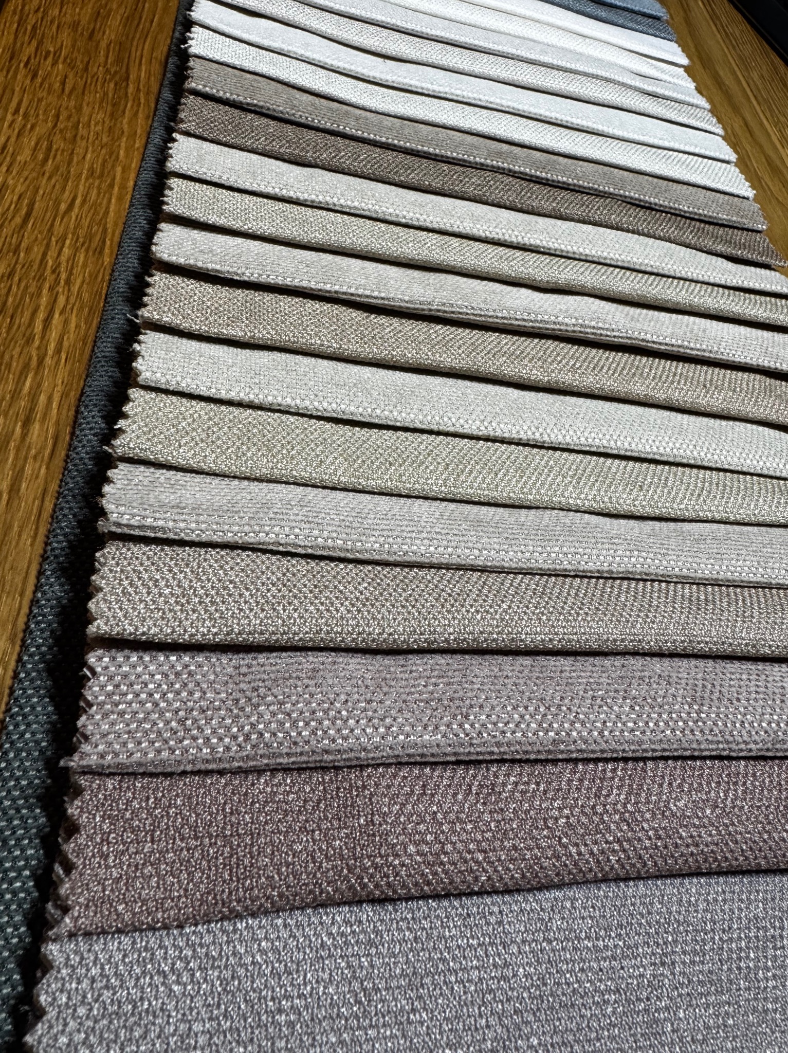 Próbki tkanin w odcieniach beżu, szarości i brązu, ułożone warstwami na drewnianym blacie. Widoczna faktura materiałów i różnorodność kolorów.