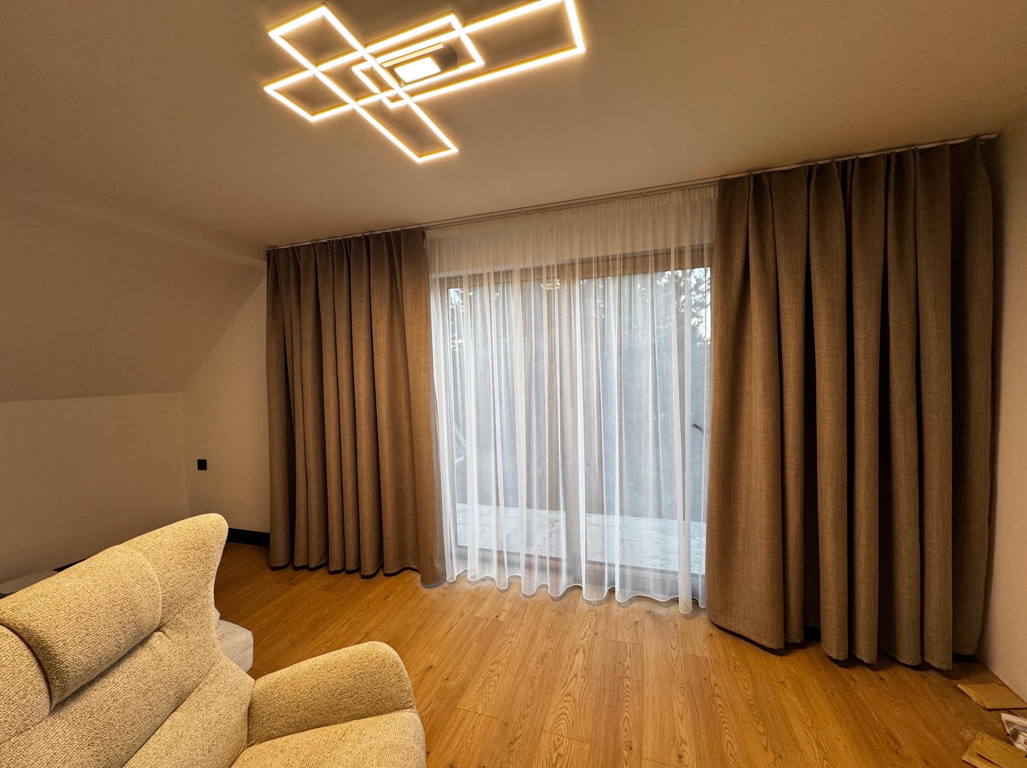 Beżowe zasłony i firanka w jasnym pokoju z drewnianą podłogą. Nad oknem karnisz. Na suficie nowoczesna lampa. Częściowo widoczny fotel w lewym dolnym rogu.