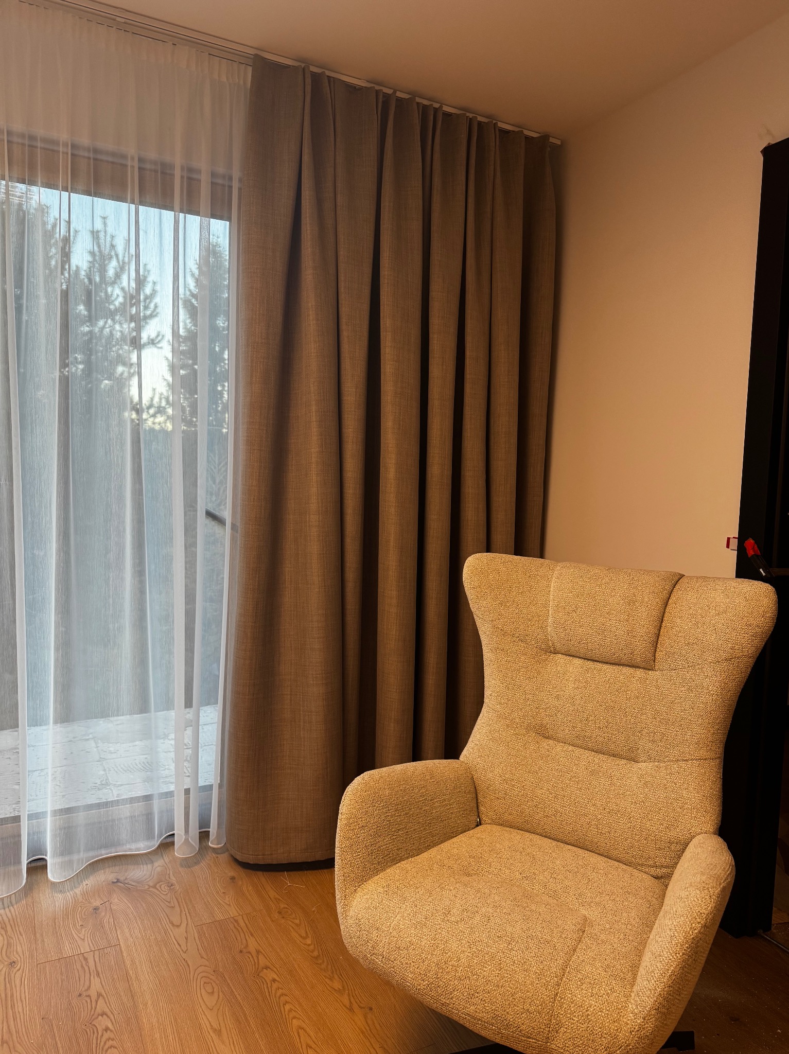 Beżowe zasłony zaciemniające i firanka w oknie, obok fotel w Rupniowie. Widok na drzewa za oknem. Przytulne wnętrze.