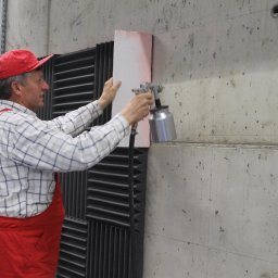 WITBUD - Pracownik w czerwonym kombinezonie maluje betonową ścianę natryskowo, używając szablonu. Widoczne panele akustyczne i surowa faktura betonu.
