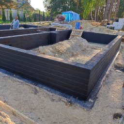 WITBUD - Wzniesione mury fundamentowe z czarnej cegły, wypełnione piaskiem, na tle zieleni drzew i częściowo widocznego domu. Widoczny pracownik budowlany i betoniarka.