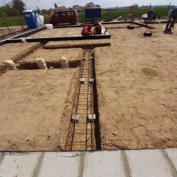 WITBUD - Zbrojenie fundamentów pod budowę domu jednorodzinnego. Widoczne szalunki, stalowe pręty i pracownicy w trakcie montażu. W tle zabudowa mieszkalna i błękitne niebo.