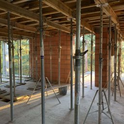 WITBUD - Wnętrze budynku w trakcie budowy: murowane ściany z pustaków, metalowe stemple podtrzymujące drewniany strop, widok na zieleń za oknem.