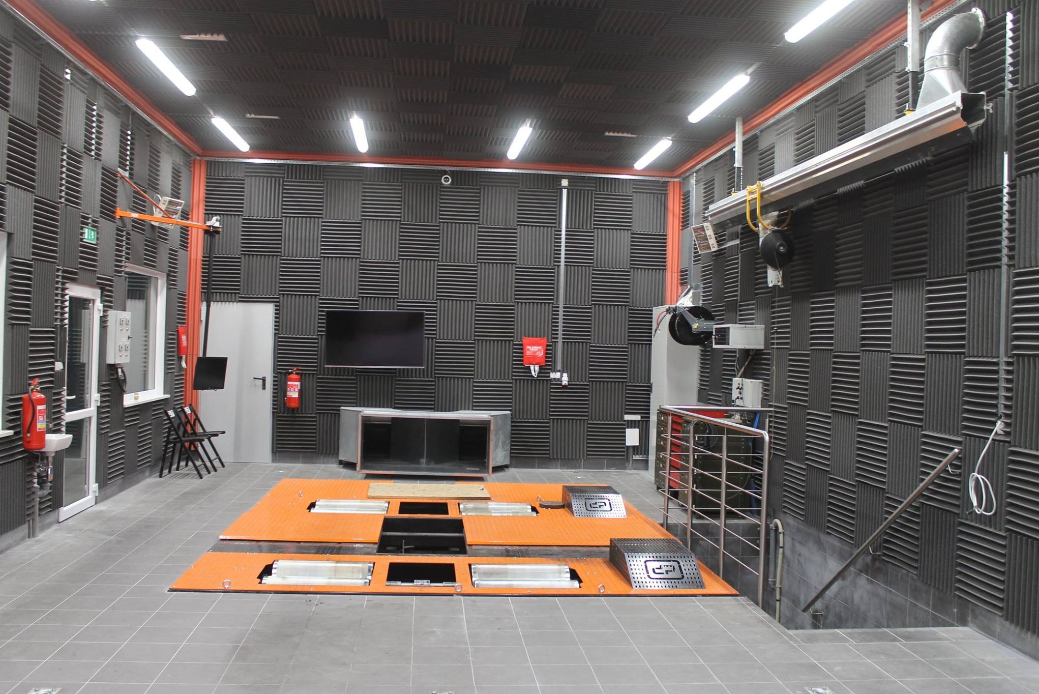 Wnętrze wyciszonego pomieszczenia z panelami akustycznymi na ścianach i suficie, pomarańczową platformą testową na środku i oświetleniem jarzeniowym.