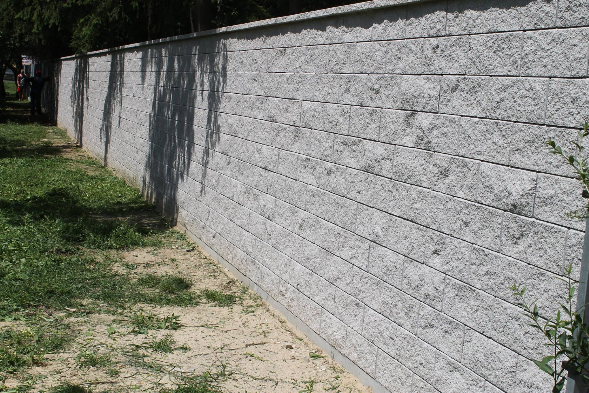 Długi, szary mur z bloków betonowych, widziany z boku, z cieniem drzewa na powierzchni i zielenią u podstawy. Widoczna faktura bloków i regularne spoiny.