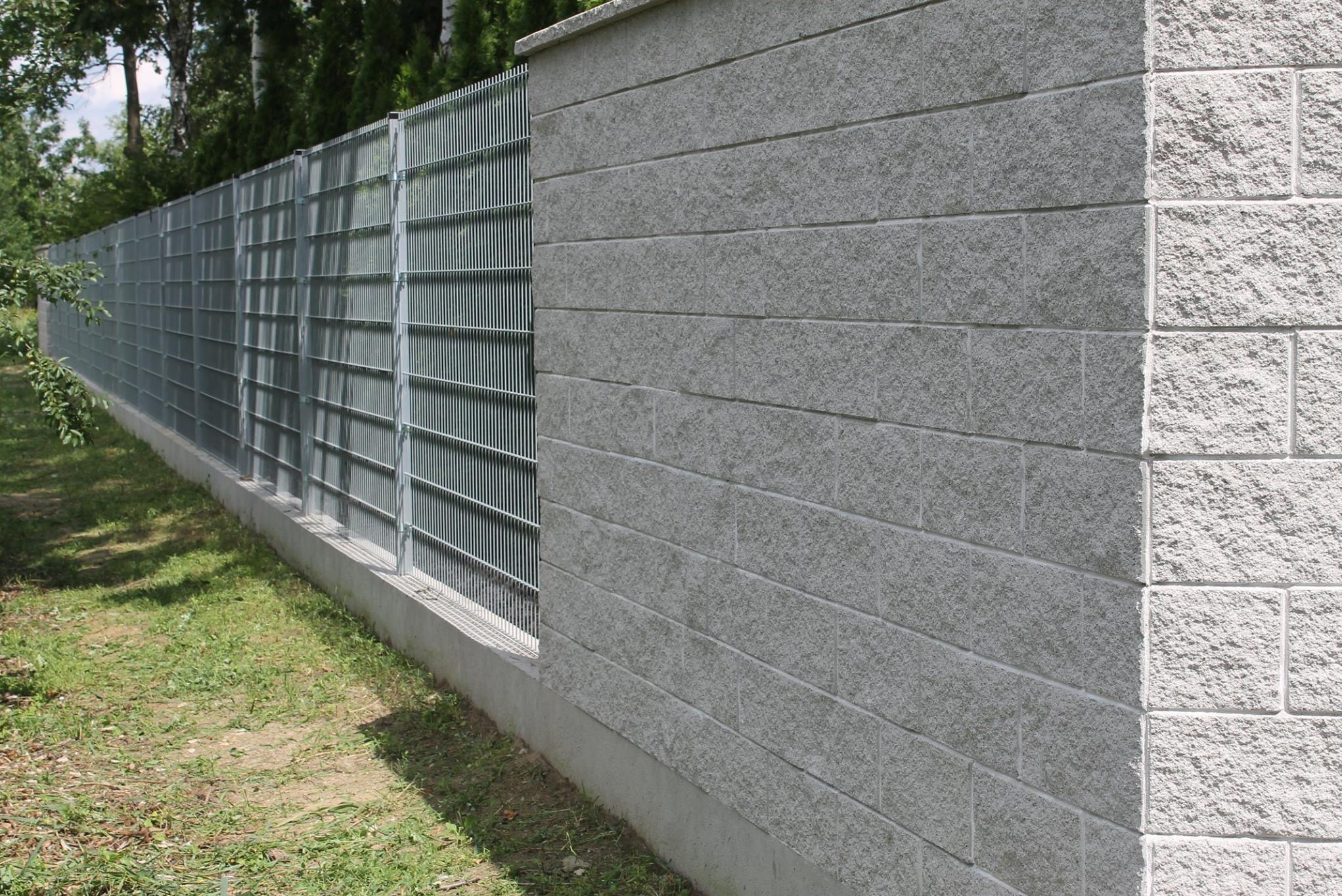 Nowoczesne ogrodzenie z szarej, teksturowanej cegły betonowej i metalowych paneli, oddzielające posesję od zieleni. Widok z poziomu gruntu, w słoneczny dzień.