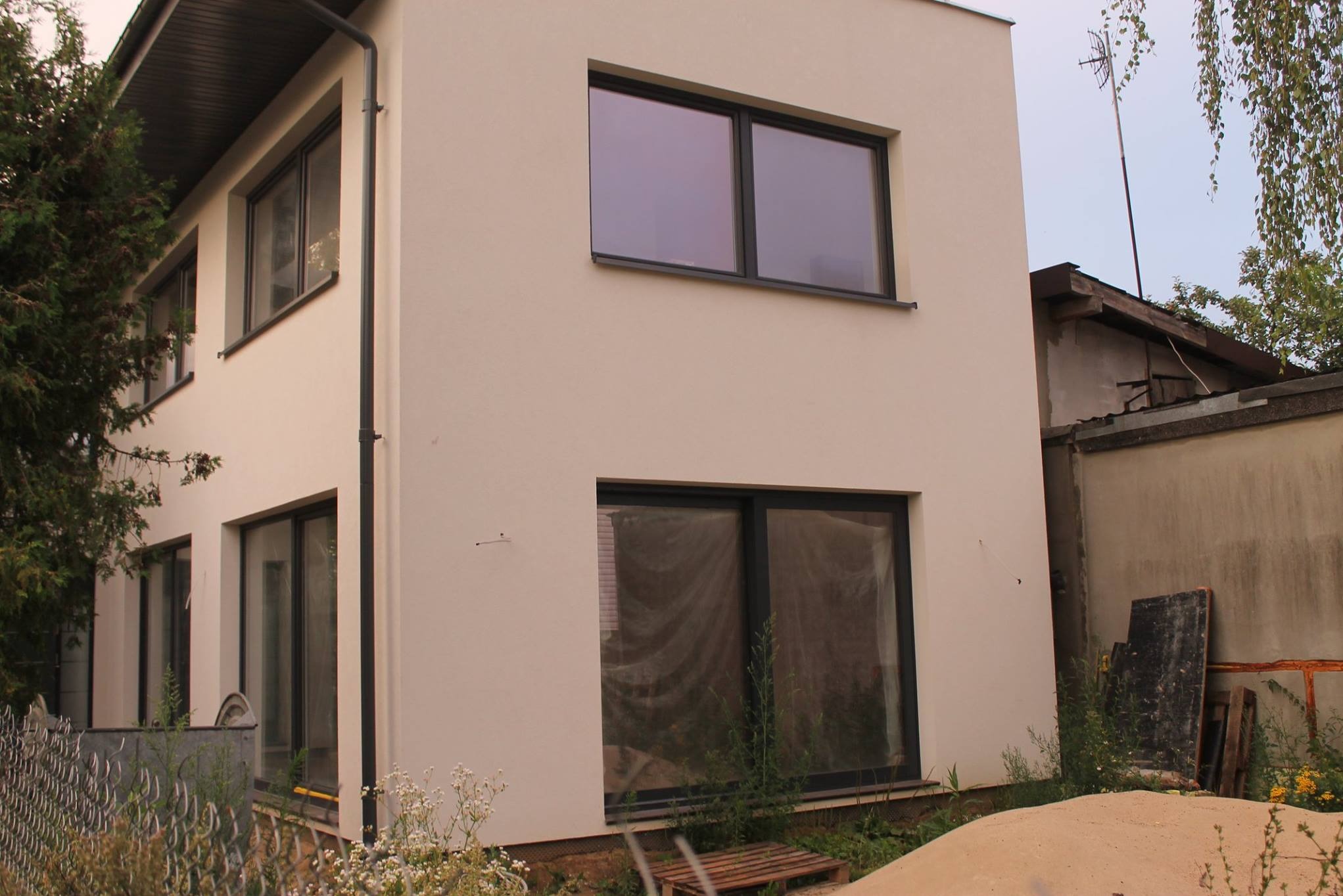 Dwukondygnacyjny dom z jasną elewacją i ciemnymi ramami okiennymi. Widoczne rośliny przed budynkiem i sąsiedni budynek w tle. Minimalistyczny styl.