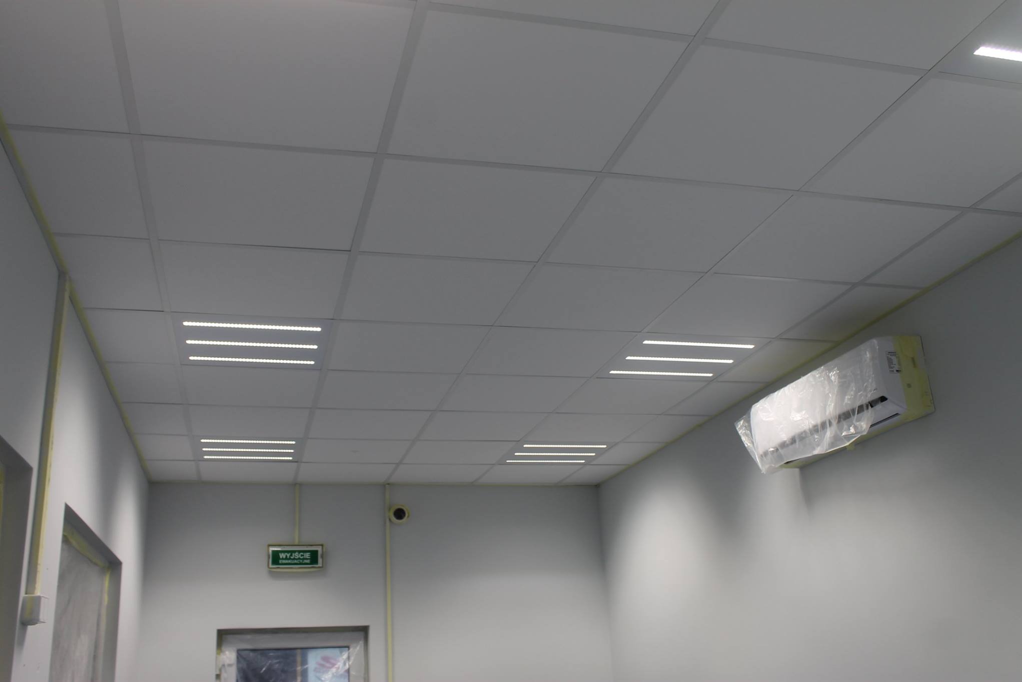 Kwadratowy sufit podwieszany z wbudowanym oświetleniem LED, klimatyzator na ścianie, widoczny znak ewakuacyjny. Minimalistyczne wnętrze biurowe.