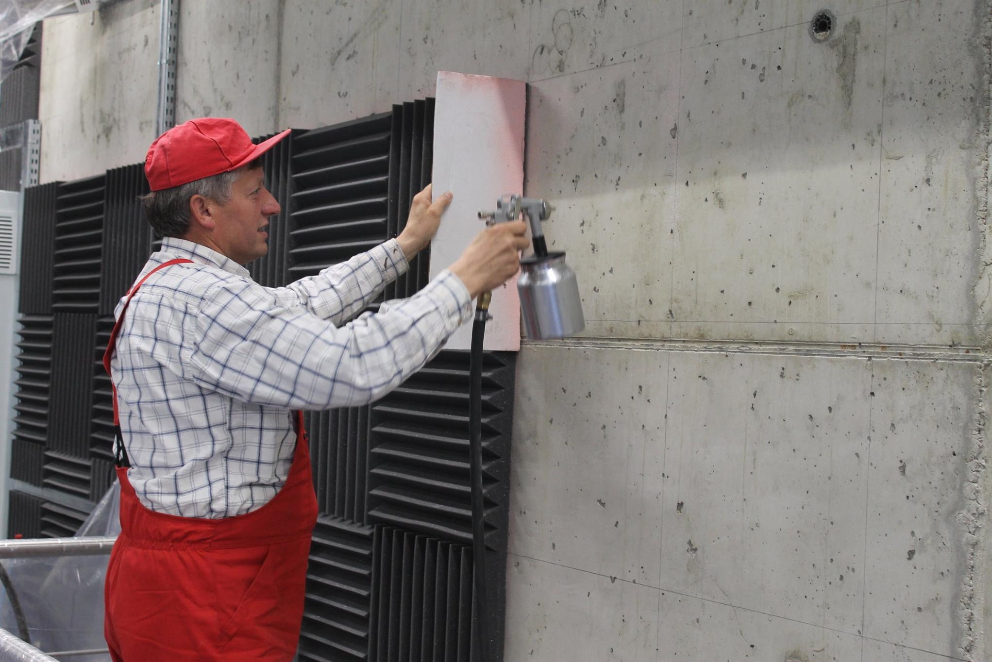 Pracownik w czerwonym kombinezonie maluje betonową ścianę natryskowo, używając szablonu. Widoczne panele akustyczne i surowa faktura betonu.