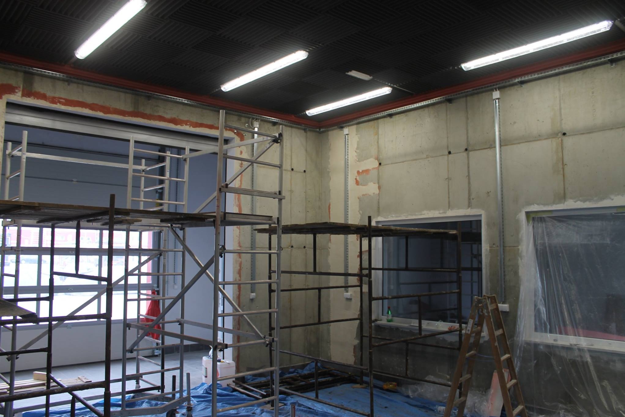 Wnętrze w trakcie prac wykończeniowych: surowy beton, rusztowania, okna zabezpieczone folią, przygotowania do malowania. Widoczne instalacje pod sufitem.