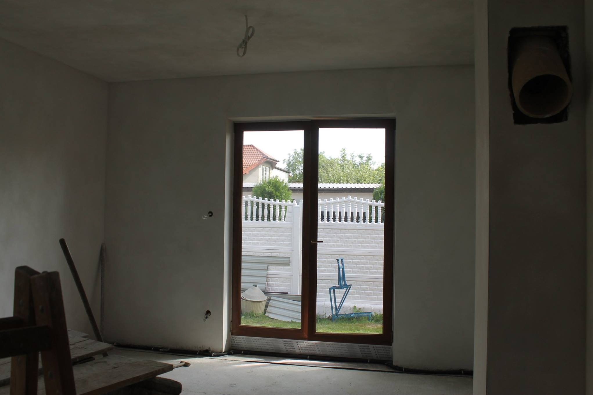 Wnętrze w stanie deweloperskim z oknem na ogród. Ściany przygotowane do malowania, widoczne kable i otwór wentylacyjny. Na podłodze wylewka.