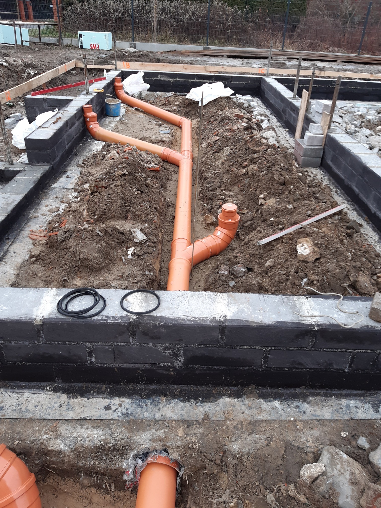 Fundamenty budynku z ułożoną instalacją kanalizacyjną z pomarańczowych rur. Widoczne czarne bloczki fundamentowe i wykop pod instalacje.