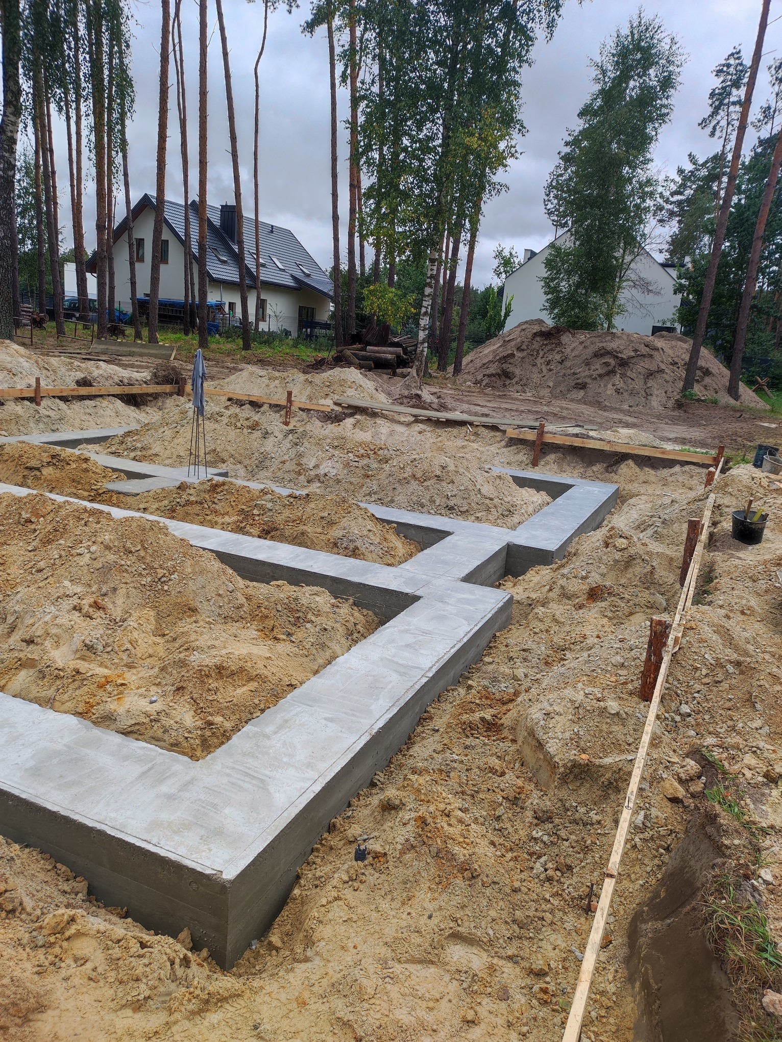 Wylane fundamenty pod dom jednorodzinny na zalesionej działce. Widoczne szalunki i betonowe ławy fundamentowe, przygotowane do dalszych prac budowlanych.