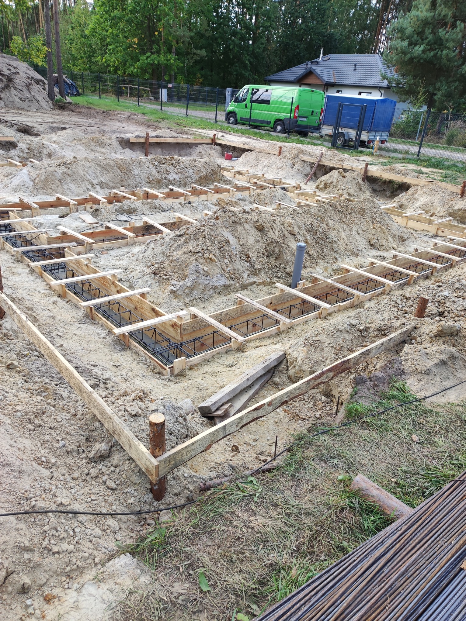 Przygotowanie szalunków i zbrojenia pod fundamenty domu jednorodzinnego na działce z zielenią w tle. Widoczne drewniane konstrukcje i stalowe pręty.