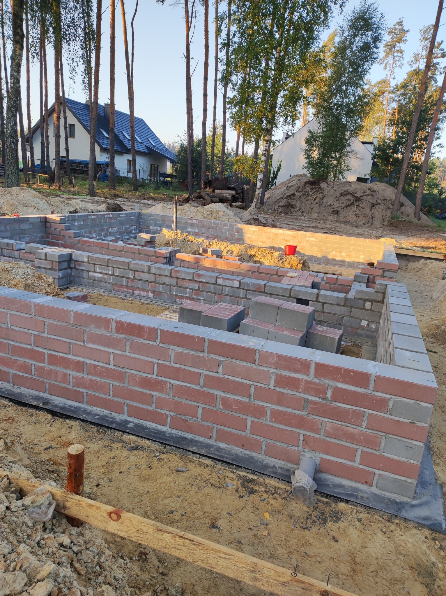 Wznoszenie fundamentów z cegły klinkierowej i bloczków betonowych na tle sosnowego lasu i domu w budowie. Widoczne detale: izolacja, rury. Ujęcie z poziomu gruntu.