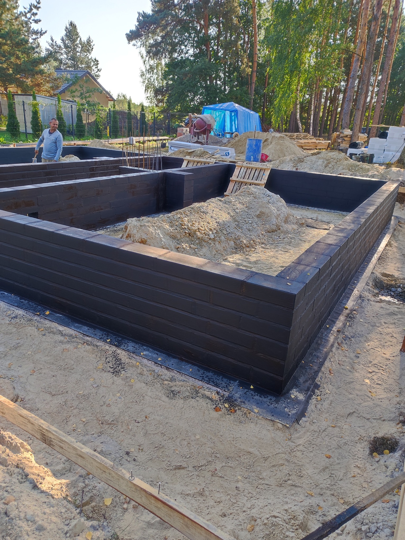 Wzniesione mury fundamentowe z czarnej cegły, wypełnione piaskiem, na tle zieleni drzew i częściowo widocznego domu. Widoczny pracownik budowlany i betoniarka.