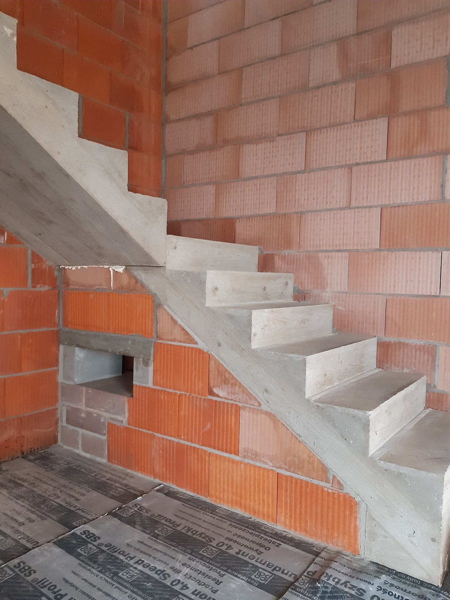 Betonowe schody w surowym stanie, w trakcie budowy, widoczne ceglane ściany i izolacja podłogi. Detal budowlany, etap prac wykończeniowych.