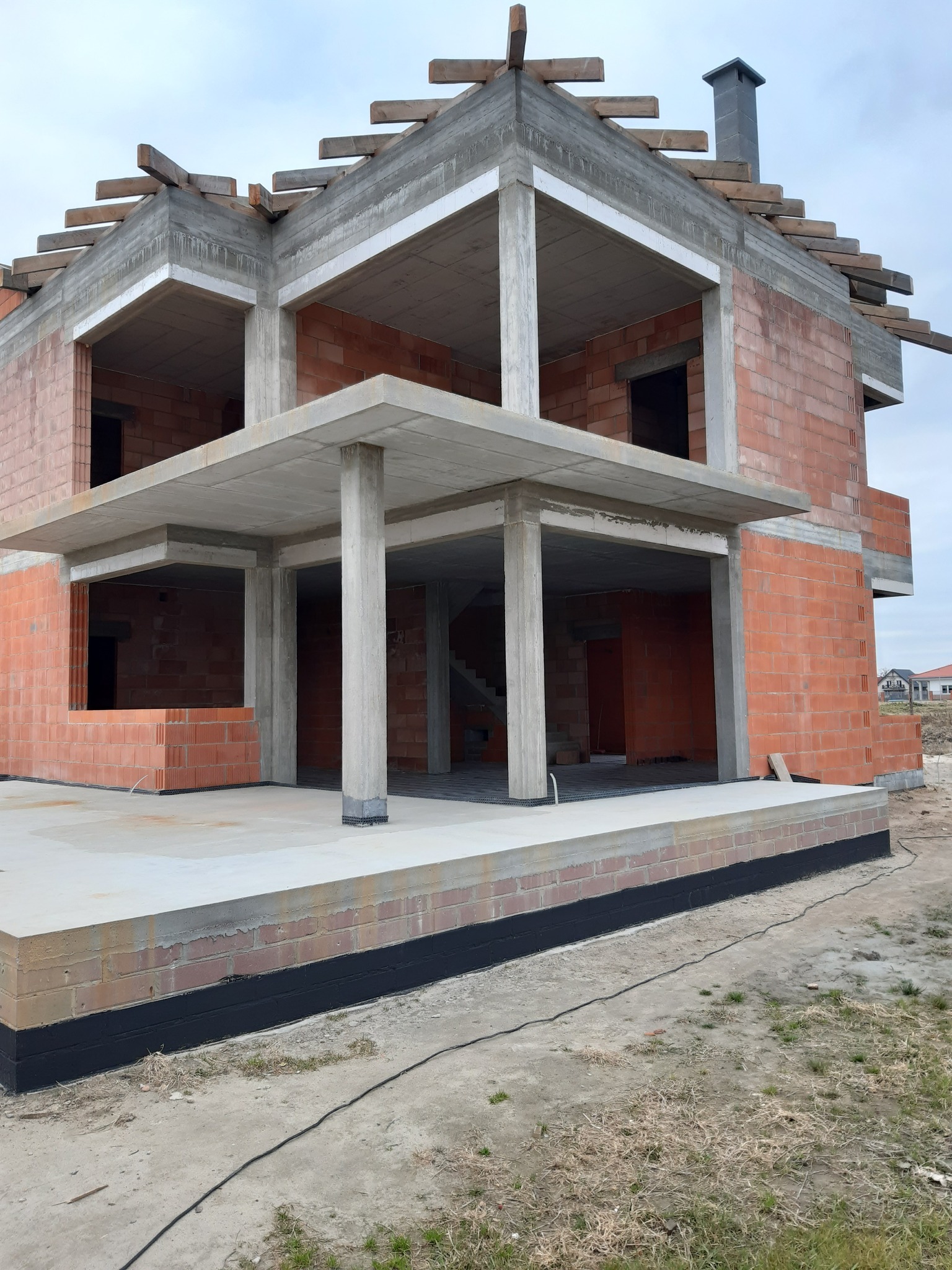 Surowy stan domu z czerwonej cegły i betonu, widoczne zbrojenia dachu i komin. Fundamenty zabezpieczone czarną izolacją, otoczenie placu budowy.