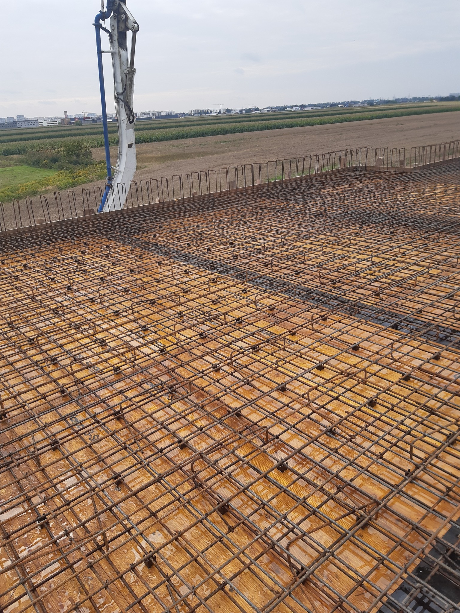 Zbrojenie stropu budynku żelbetowego z widoczną pompą do betonu w tle, ukazane z perspektywy góry. Widoczne pola uprawne w oddali.
