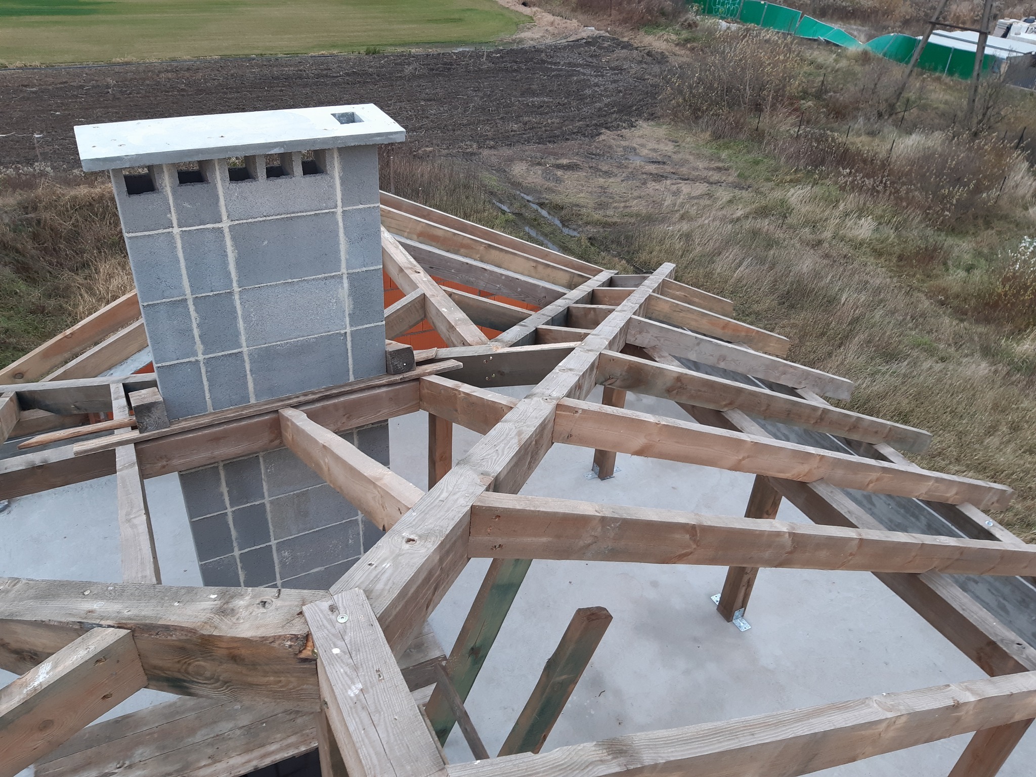 Wysoki kąt: Konstrukcja dachu z drewnianych belek i komin z szarej cegły na tle jesiennego krajobrazu. Widoczne elementy mocujące i surowy stan budowy.