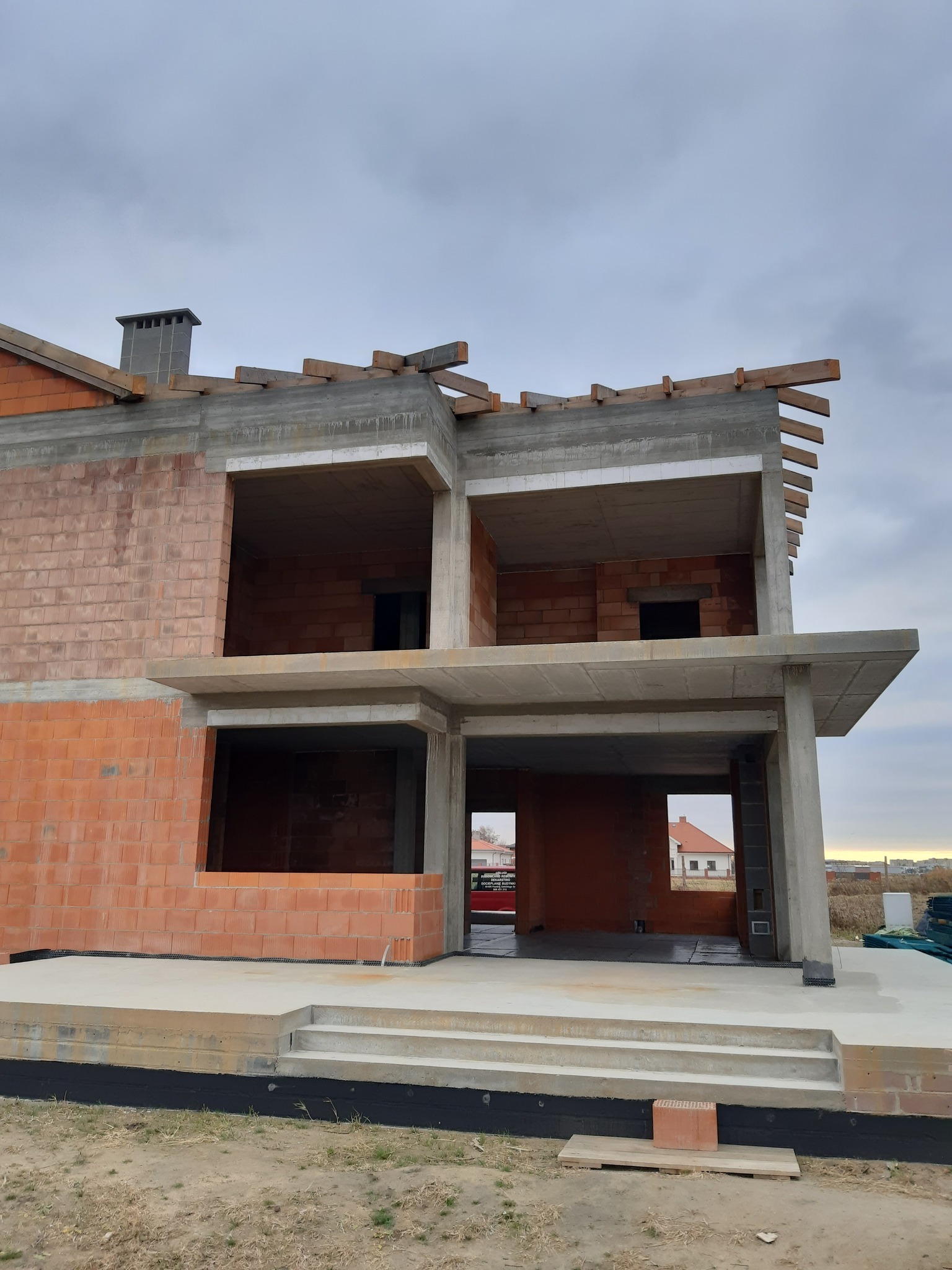 Surowy stan dwukondygnacyjnego domu z czerwonej cegły i betonu, widok z dołu, odsłonięte drewniane elementy dachu, w tle pochmurne niebo.