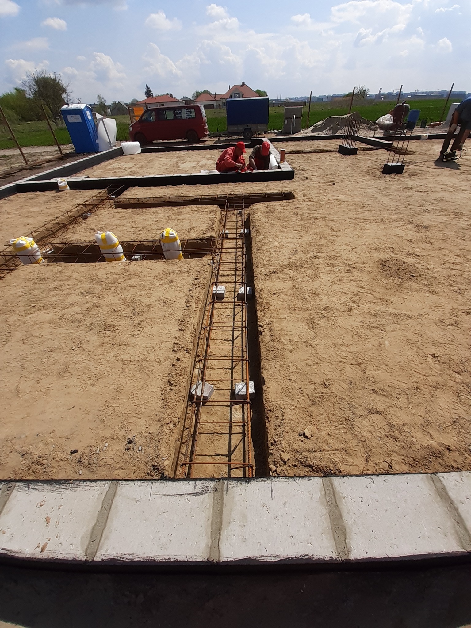 Zbrojenie fundamentów pod budowę domu jednorodzinnego. Widoczne szalunki, stalowe pręty i pracownicy w trakcie montażu. W tle zabudowa mieszkalna i błękitne niebo.