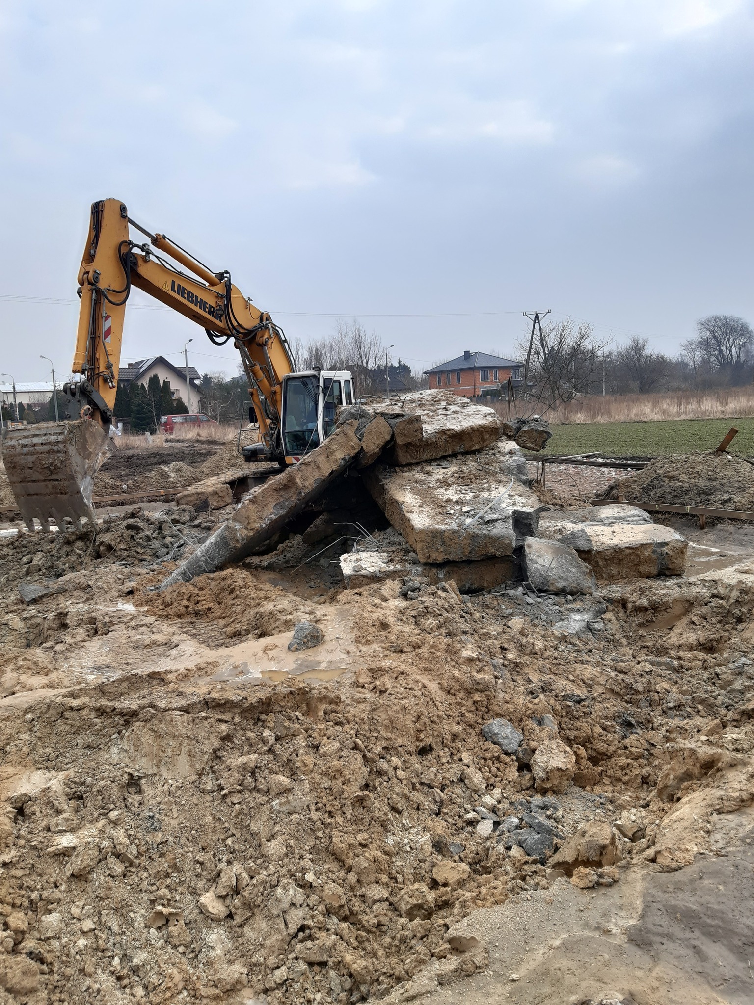 Żółta koparka Liebherr na placu budowy, gruz betonowy, ziemia. Prace ziemne w toku, widoczne domy w tle. Pochmurne niebo.