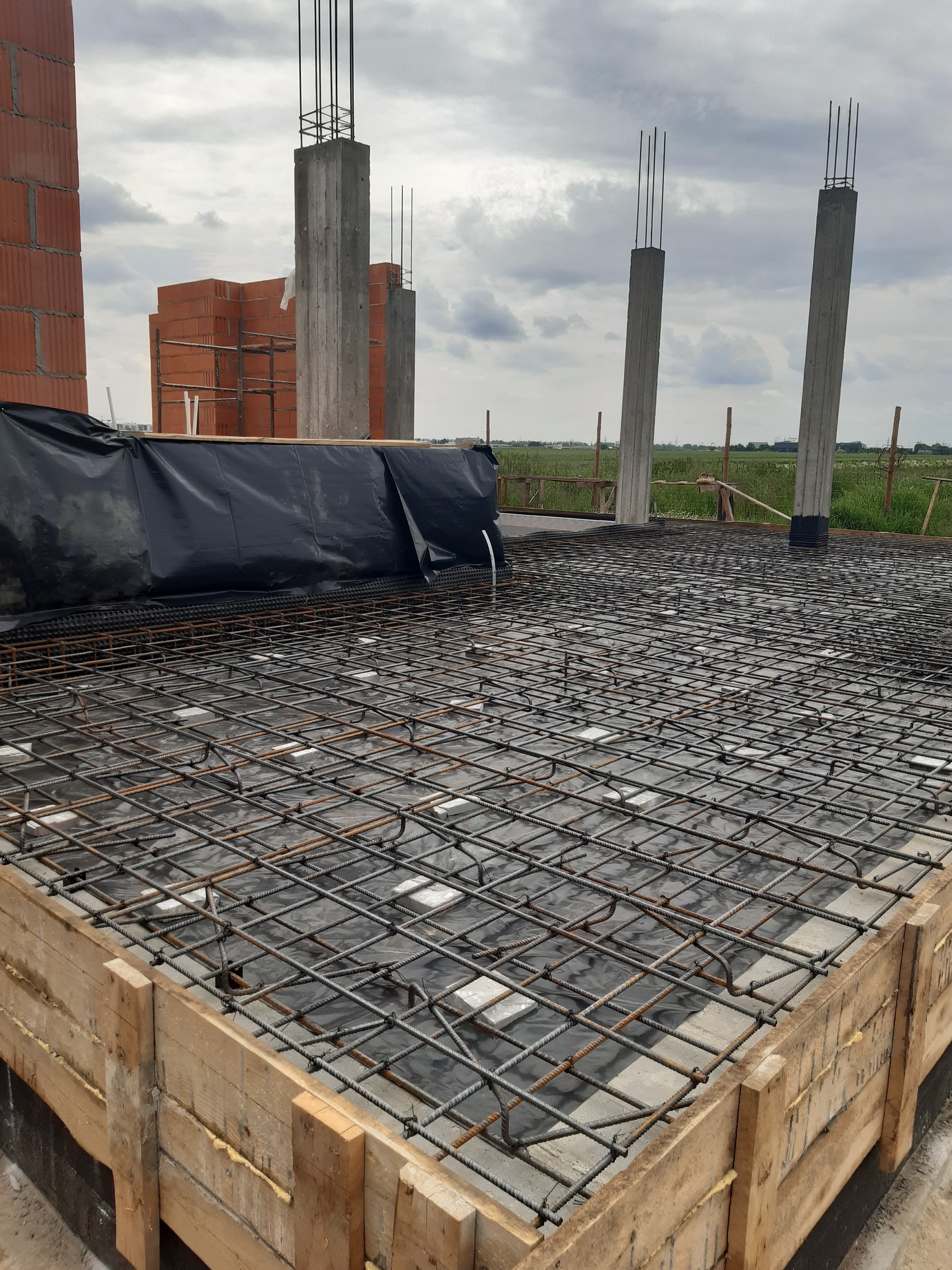 Zbrojenie fundamentu z widoczną siatką stalową, szalunkiem drewnianym i wystającymi słupami żelbetowymi. W tle cegły i zielona łąka pod pochmurnym niebem.
