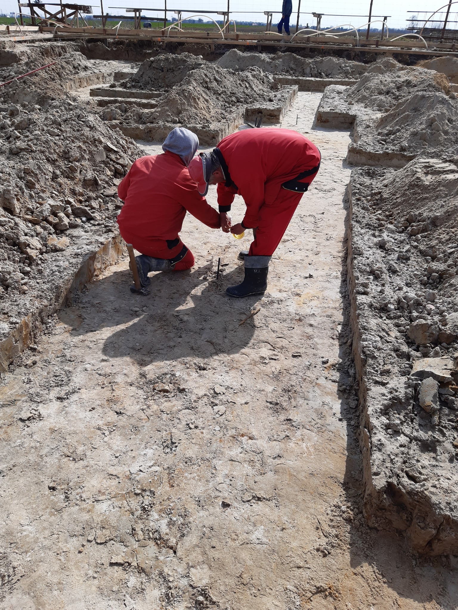 Dwóch robotników w czerwonych kombinezonach pracuje przy wykopie fundamentów, używając narzędzi do precyzyjnego ustawiania. W tle widoczne drewniane konstrukcje.