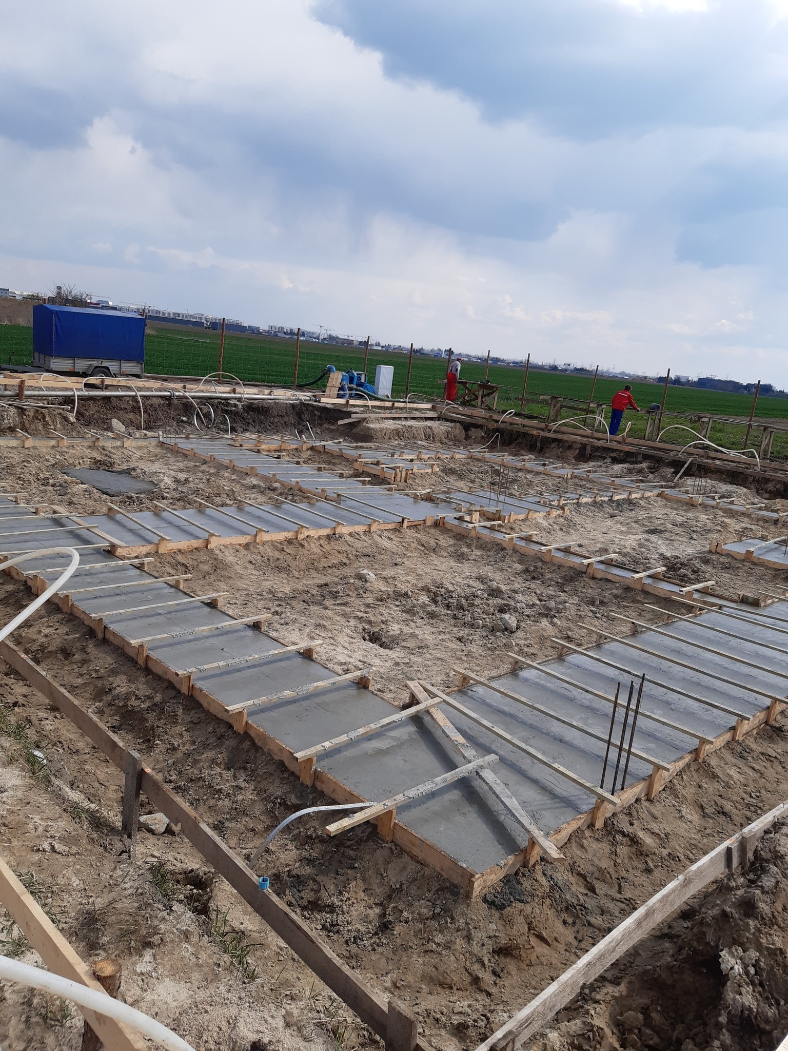 Wylane fundamenty pod budowę domu jednorodzinnego. Widoczne szalunki wypełnione betonem, teren budowy, pracownicy w oddali i błękitne niebo z chmurami.