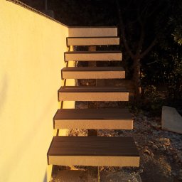 Dream Works Renovations - Zewnętrzne schody z drewnopodobnych desek prowadzące w górę, przylegające do jasnej ściany. Widoczne elementy konstrukcyjne i otoczenie w naturalnym świetle.