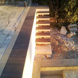 Dream Works Renovations - Wykończenie schodów zewnętrznych z drewnopodobnych desek kompozytowych, widok z góry na fragment tarasu z kamienną posadzką i odpływem liniowym. Prace w toku.