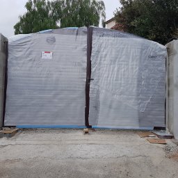 Dream Works Renovations - Nowa, brązowa brama wjazdowa, zabezpieczona folią ochronną, osadzona w surowych, betonowych słupkach. Widoczne elementy montażowe i świeży beton.