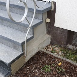 Dream Works Renovations - Granitowe schody zewnętrzne z metalową balustradą, prowadzące do budynku z białą elewacją i brązowym cokołem. Widoczna kratka wentylacyjna i rabata z roślinami.