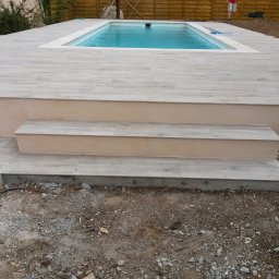 Dream Works Renovations - Wykończony taras z jasnoszarej płytki wokół basenu, z niskimi schodami wejściowymi. Widoczne niedokończone prace ziemne wokół tarasu.