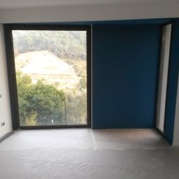 Dream Works Renovations - Pokój po malowaniu: ściana w odcieniu głębokiego granatu kontrastuje z bielą, okno z widokiem na zieleń, podłoga zabezpieczona folią malarską. Minimalistyczne gniazdka na ścianie.