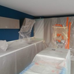 Dream Works Renovations - Kuchnia w trakcie zabezpieczania folią malarską przed malowaniem ścian na granatowo. Widoczne szafki i blaty zakryte folią ochronną. Przygotowania do prac malarskich.