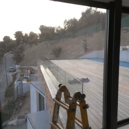 Dream Works Renovations - Nowoczesny taras z drewnianą podłogą i szklaną balustradą, widok z okna. Na pierwszym planie drabina, w tle basen i zieleń wzgórza.