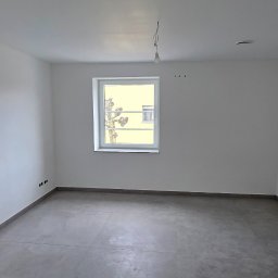 Dream Works Renovations - Pusty, odnowiony pokój z szarymi płytkami podłogowymi i białymi ścianami. Widoczne okno z widokiem na drzewo i fragment białych drzwi po prawej stronie kadru.