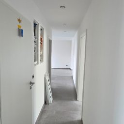 Dream Works Renovations - Długi, jasny korytarz z szarą podłogą w nowym budynku. Widoczne białe ściany, drzwi i instalacje elektryczne w trakcie montażu. Minimalistyczny styl wykończenia.