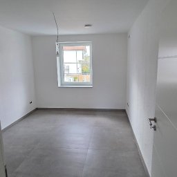 Dream Works Renovations - Pusty pokój po remoncie z szarą podłogą z płytek, białymi ścianami i drzwiami. Widoczne okno z widokiem na zewnątrz. Minimalistyczne wykończenie wnętrza.