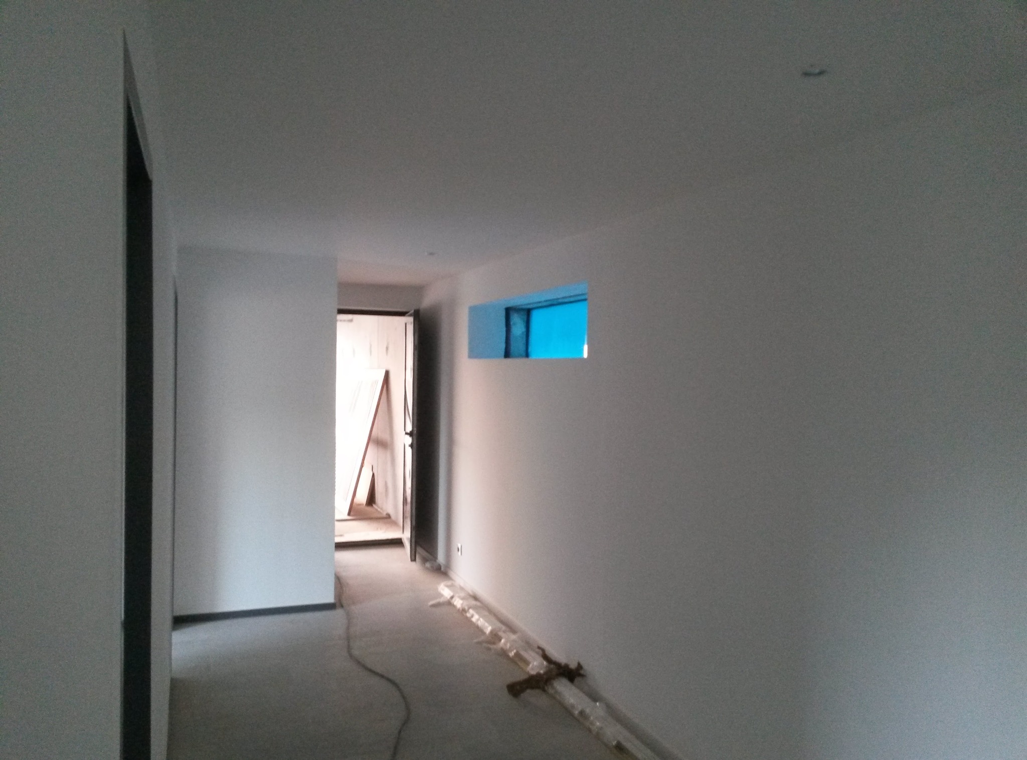 Białe ściany korytarza po malowaniu, widoczne małe okno z niebieskim tłem, drzwi na końcu korytarza i listwy przypodłogowe w folii na podłodze.