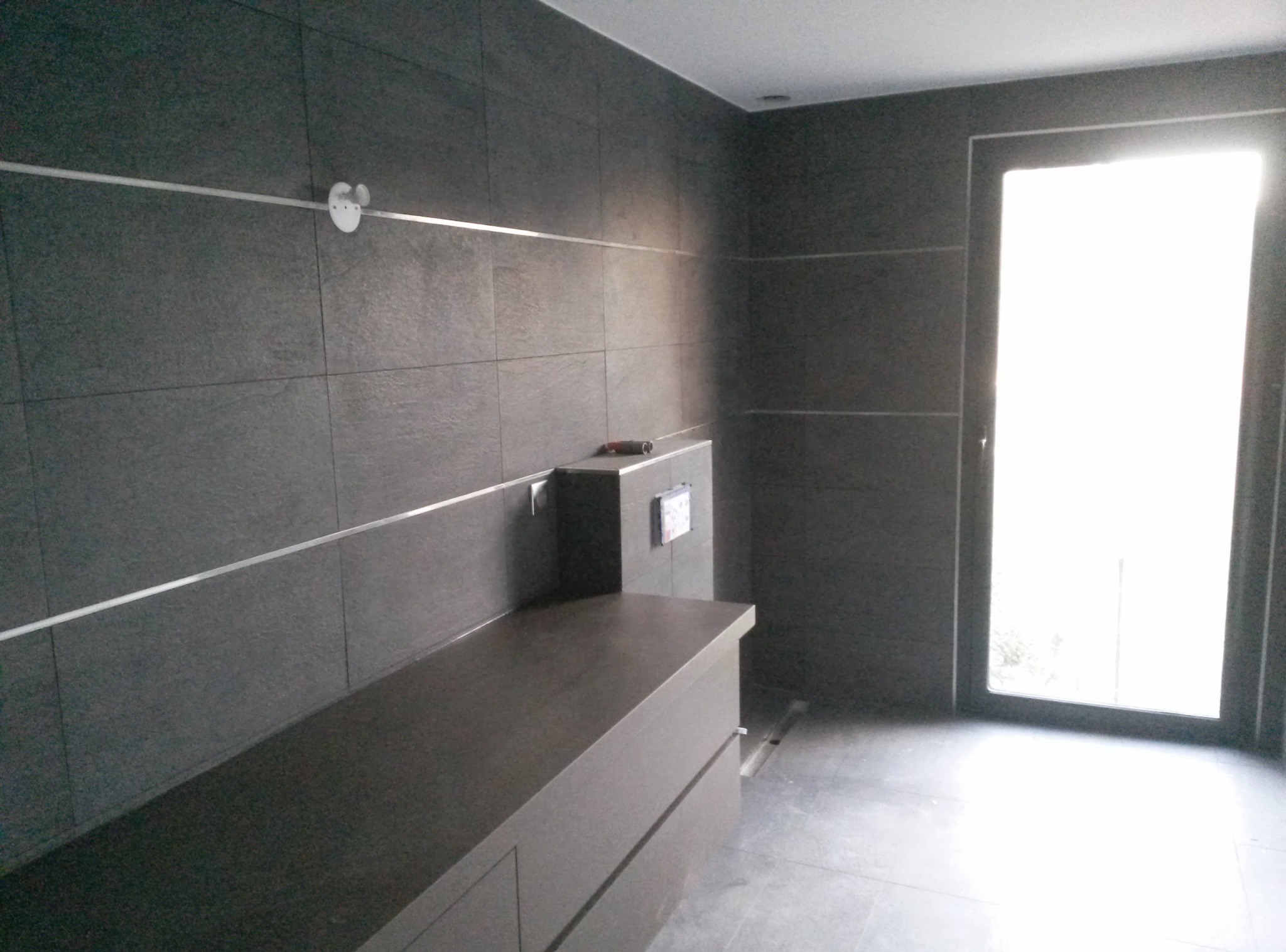 Nowoczesna łazienka z ciemnymi płytkami i minimalistycznym blatem. Widoczne srebrne listwy dekoracyjne i odpływ liniowy. Duże okno wpuszcza jasne światło.