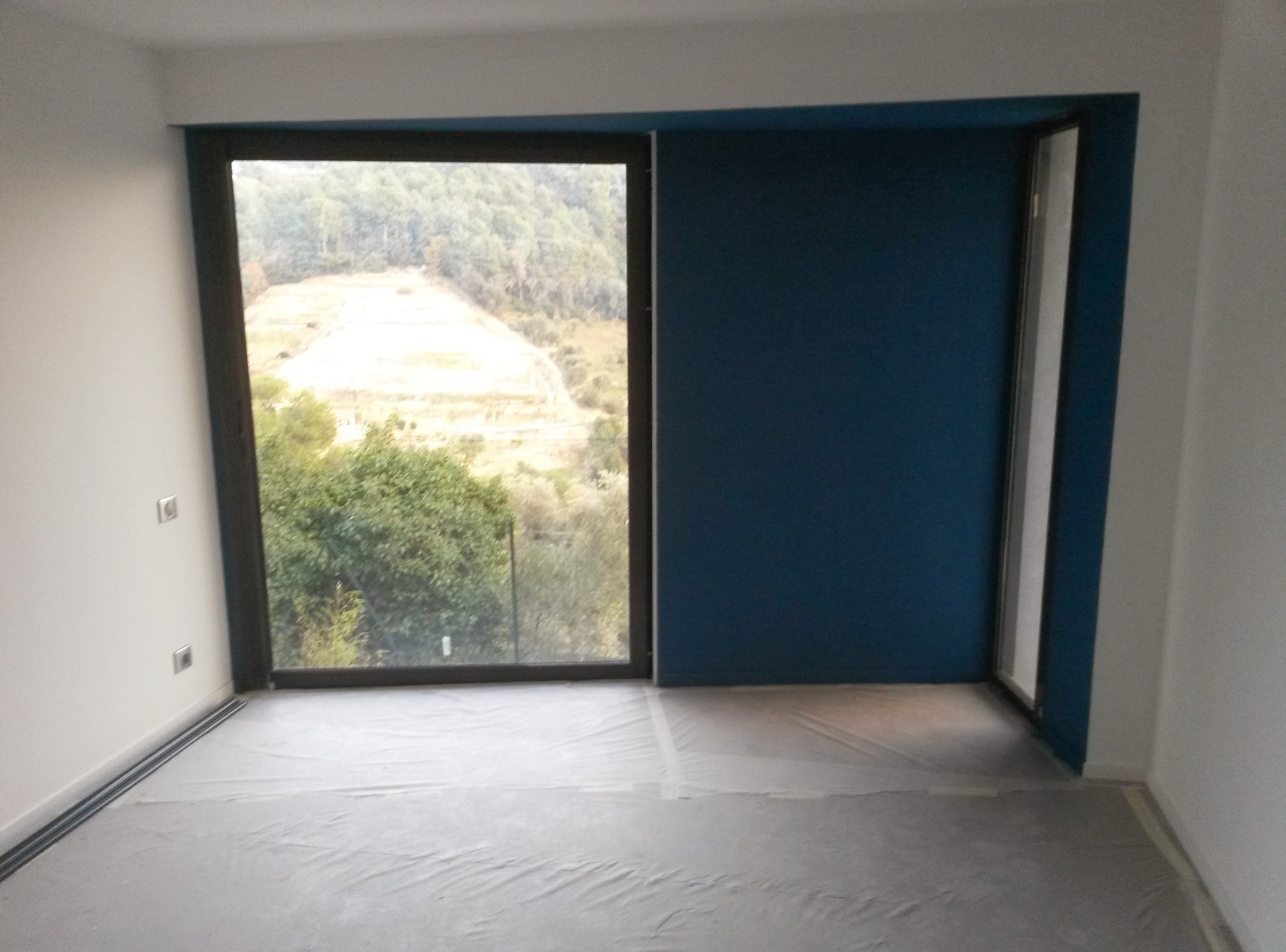 Pokój po malowaniu: ściana w odcieniu głębokiego granatu kontrastuje z bielą, okno z widokiem na zieleń, podłoga zabezpieczona folią malarską. Minimalistyczne gniazdka na ścianie.